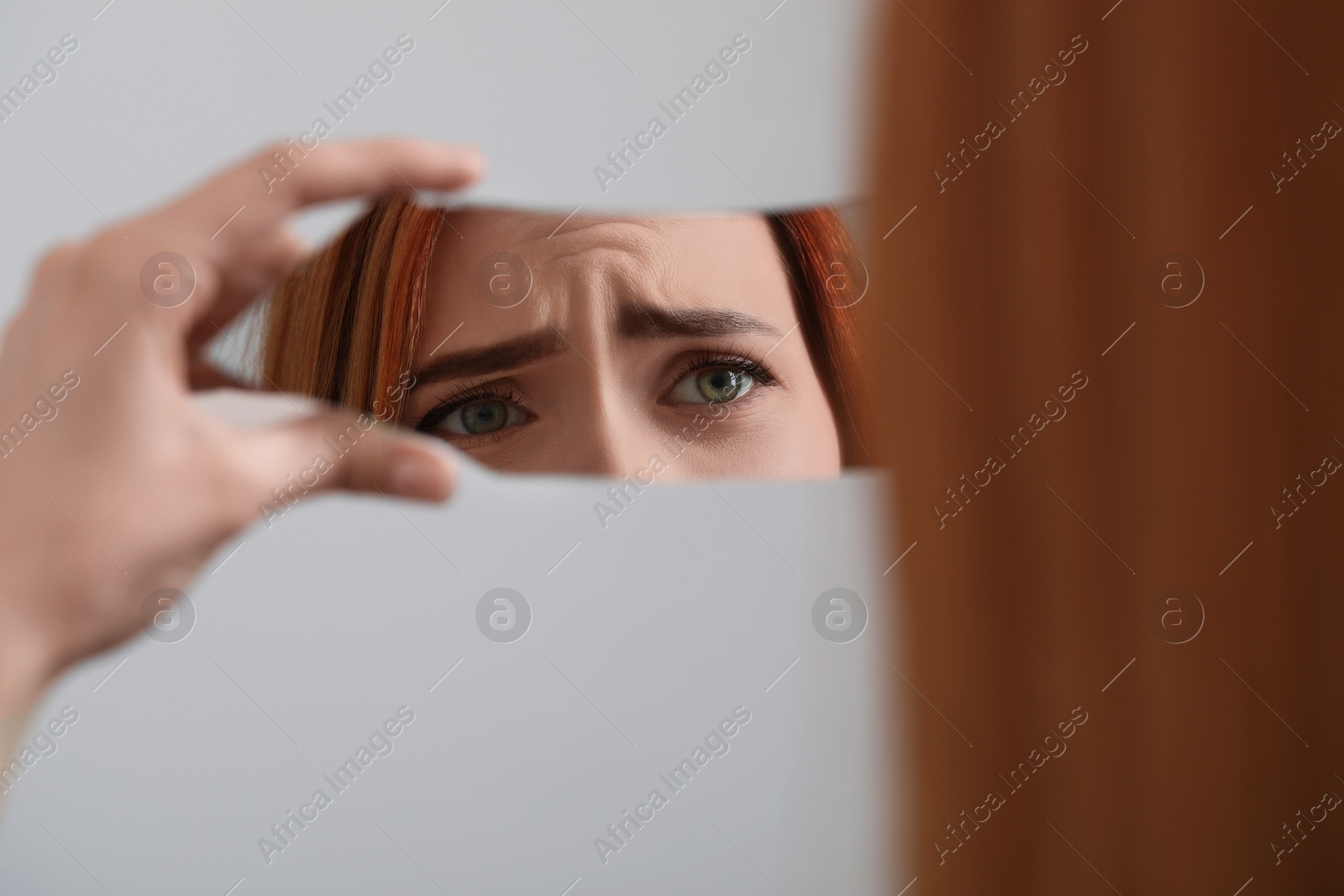 Photo of Young woman looking at herself in shard of broken mirror on light grey background, closeup