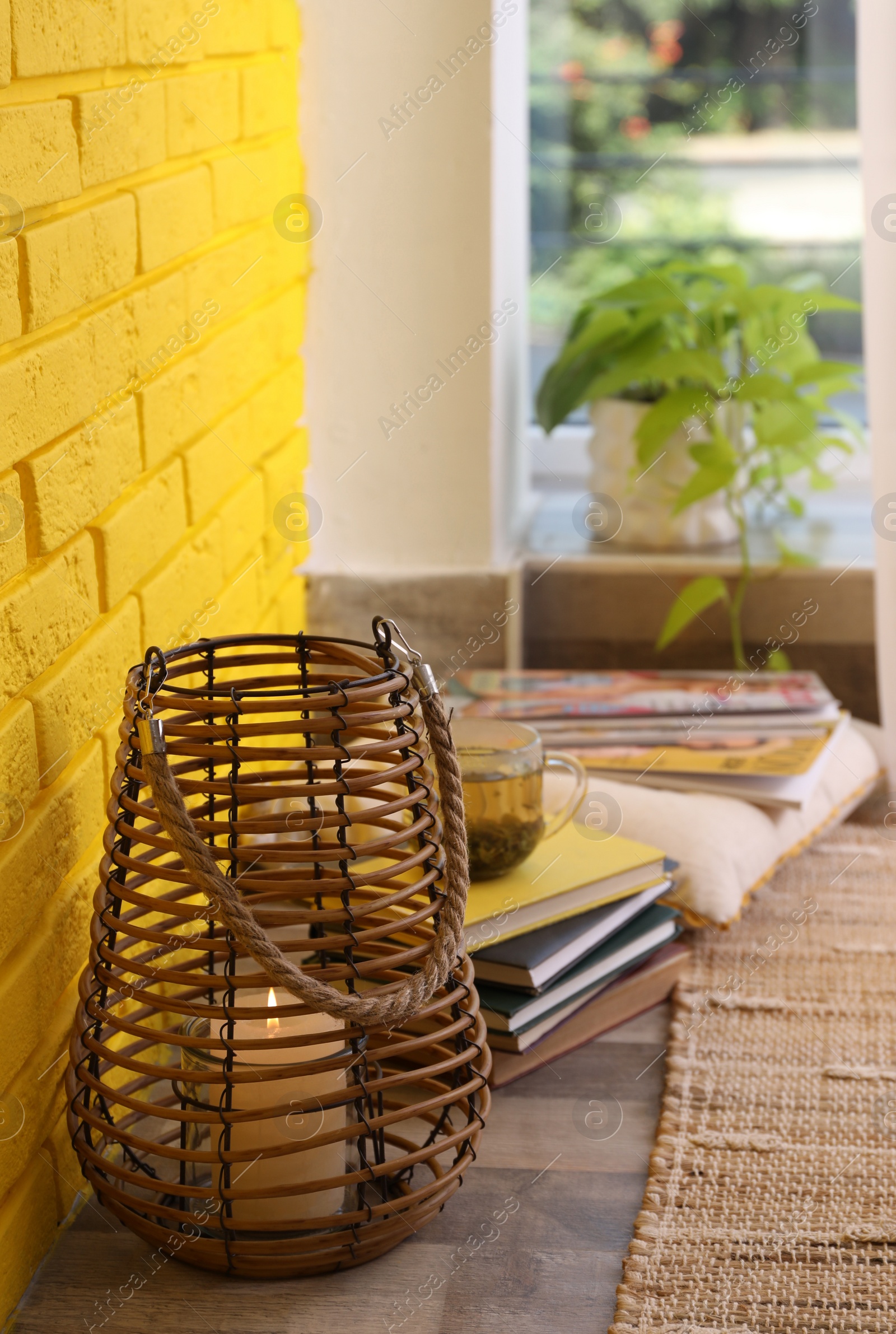 Photo of Stylish holder with burning candle and books on floor near yellow brick wall