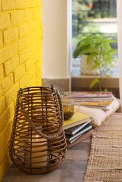 Photo of Stylish holder with burning candle and books on floor near yellow brick wall