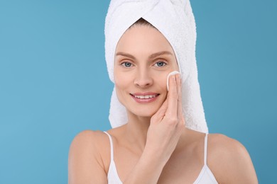 Photo of Beautiful woman in terry towel removing makeup with cotton pad on light blue background