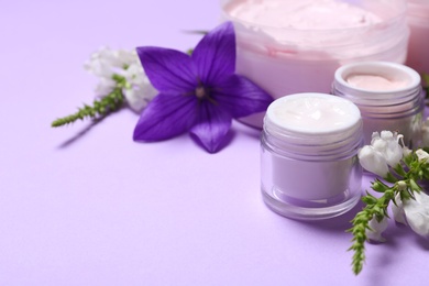 Jars of body cream and flowers on lilac background. Space for text