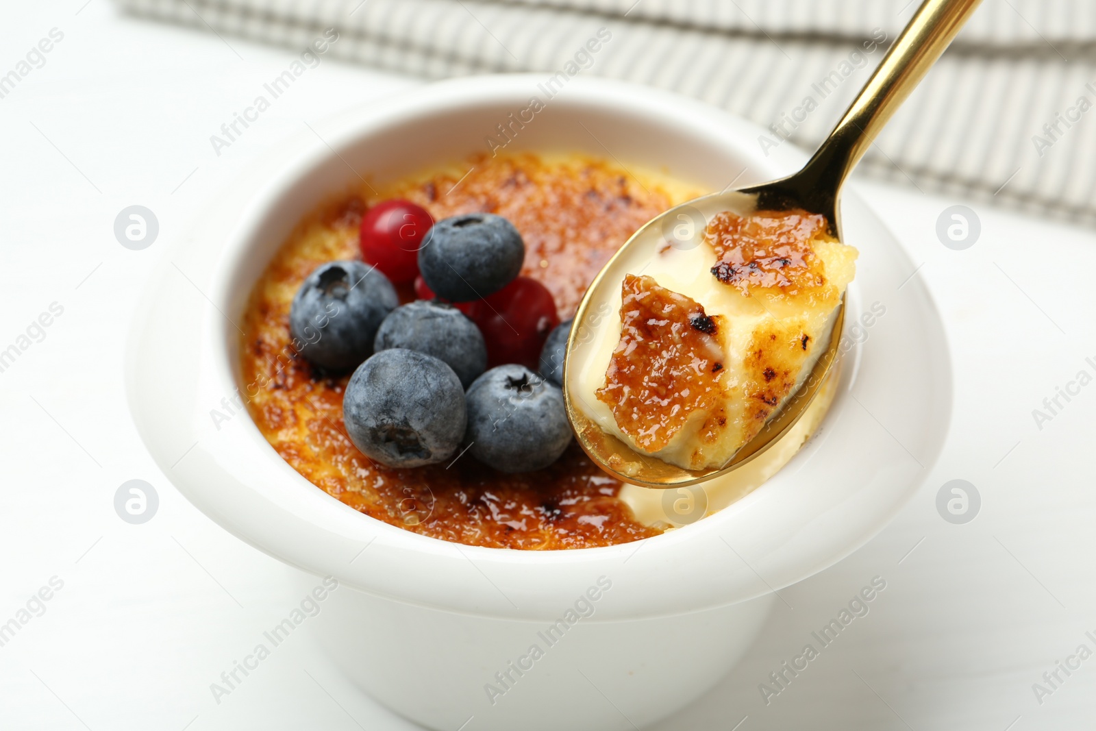 Photo of Eating delicious creme brulee with berries from spoon at white table, closeup