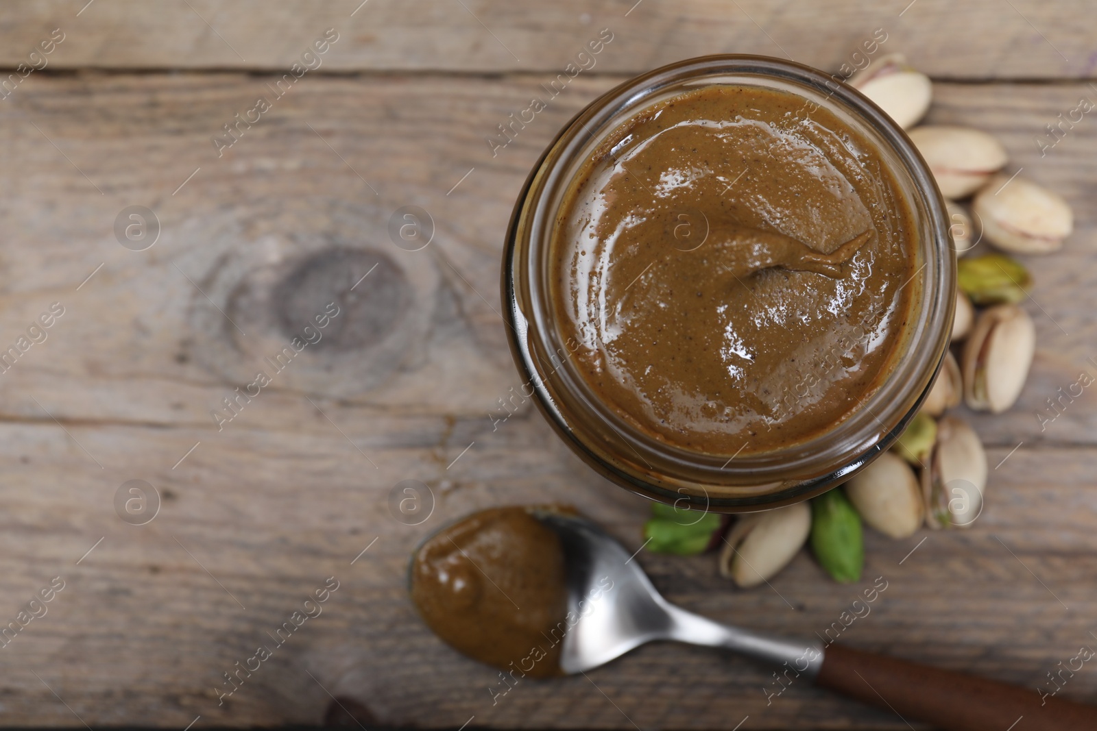 Photo of Tasty pistachio nut paste in jar and spoon on wooden table, flat lay. Space for text