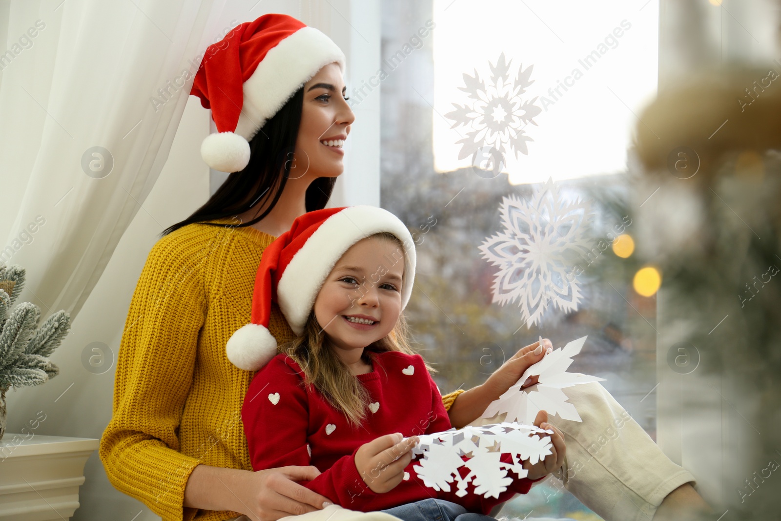 Photo of Mother and daughter in Santa hats with paper snowflakes near window at home