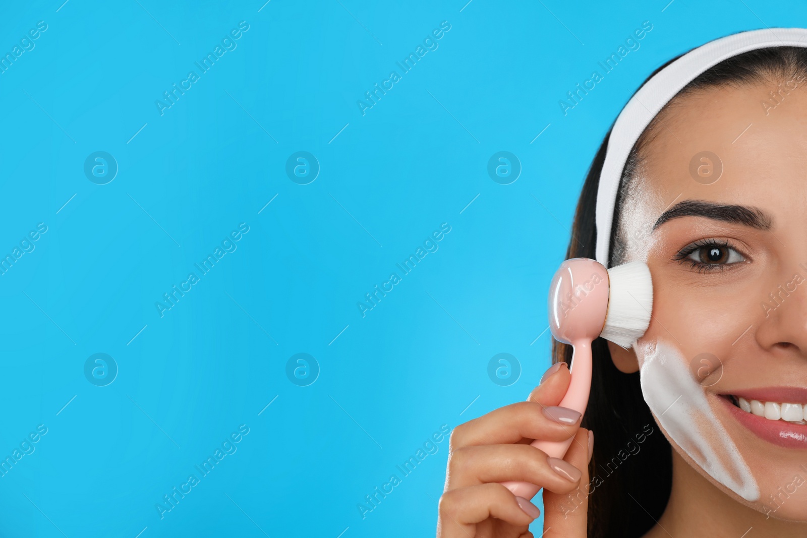 Photo of Young woman using facial cleansing brush on light blue background, space for text. Washing accessory