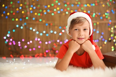 Photo of Cute little child in Santa hat lying on floor against blurred lights. Christmas celebration