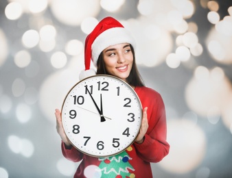 New Year countdown. Happy woman in Santa hat holding clock against blurred lights on background