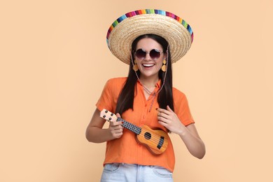 Young woman in Mexican sombrero hat playing ukulele on beige background