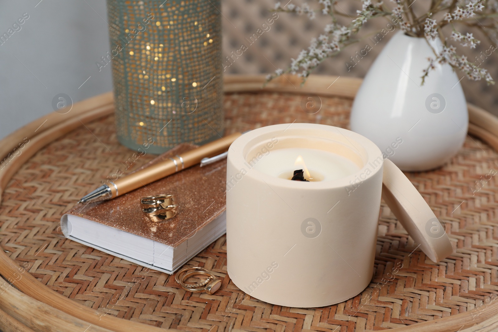 Photo of Burning soy candle, notebook and stylish accessories on wicker table indoors