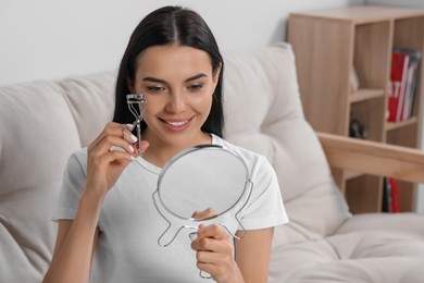 Photo of Beautiful young woman with mirror using eyelash curler indoors