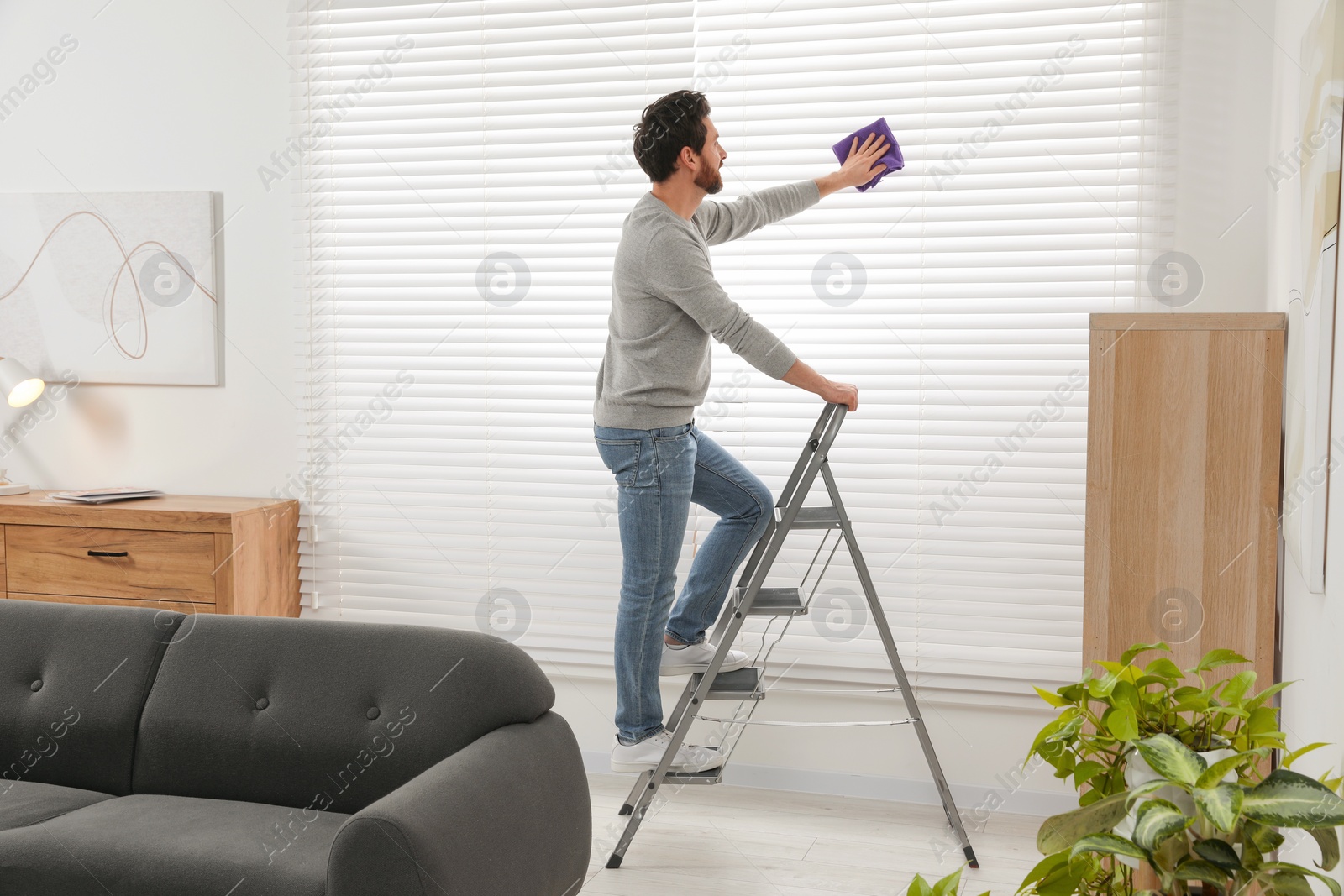 Photo of Man on metal ladder wiping blinds at home