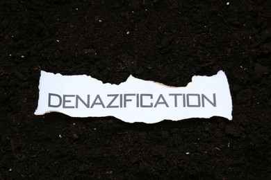 Photo of Piece of torn paper with word Denazification on soil, top view