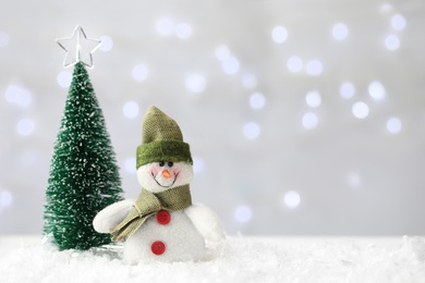 Photo of Snowman toy and Christmas tree on snow against blurred festive lights. Space for text
