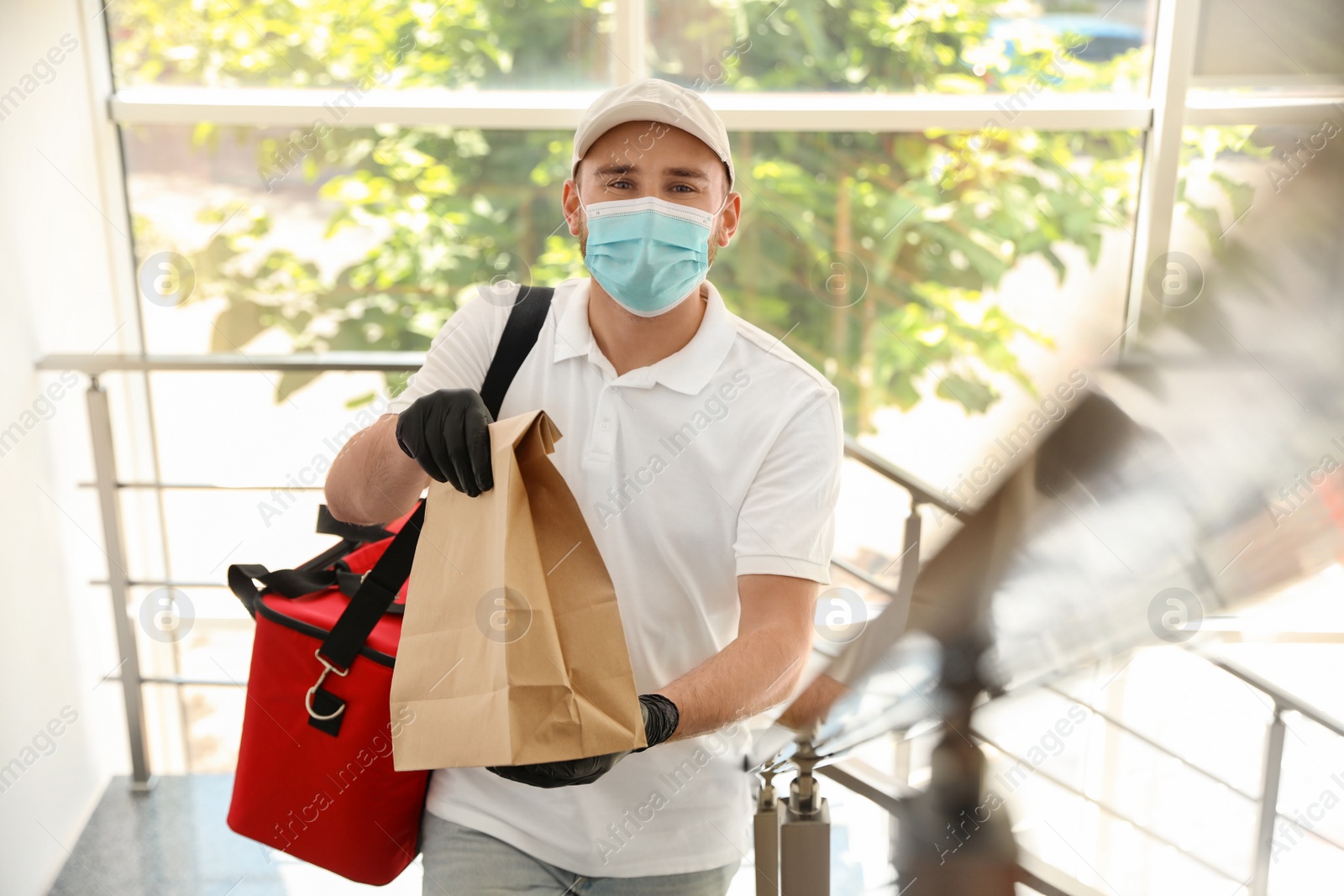 Photo of Courier in protective mask and gloves with order indoors. Restaurant delivery service during coronavirus quarantine