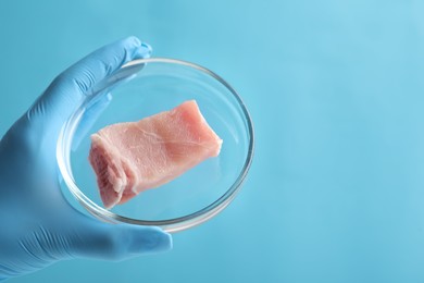 Photo of Scientist holding Petri dish with lab grown meat on light blue background, closeup. Space for text