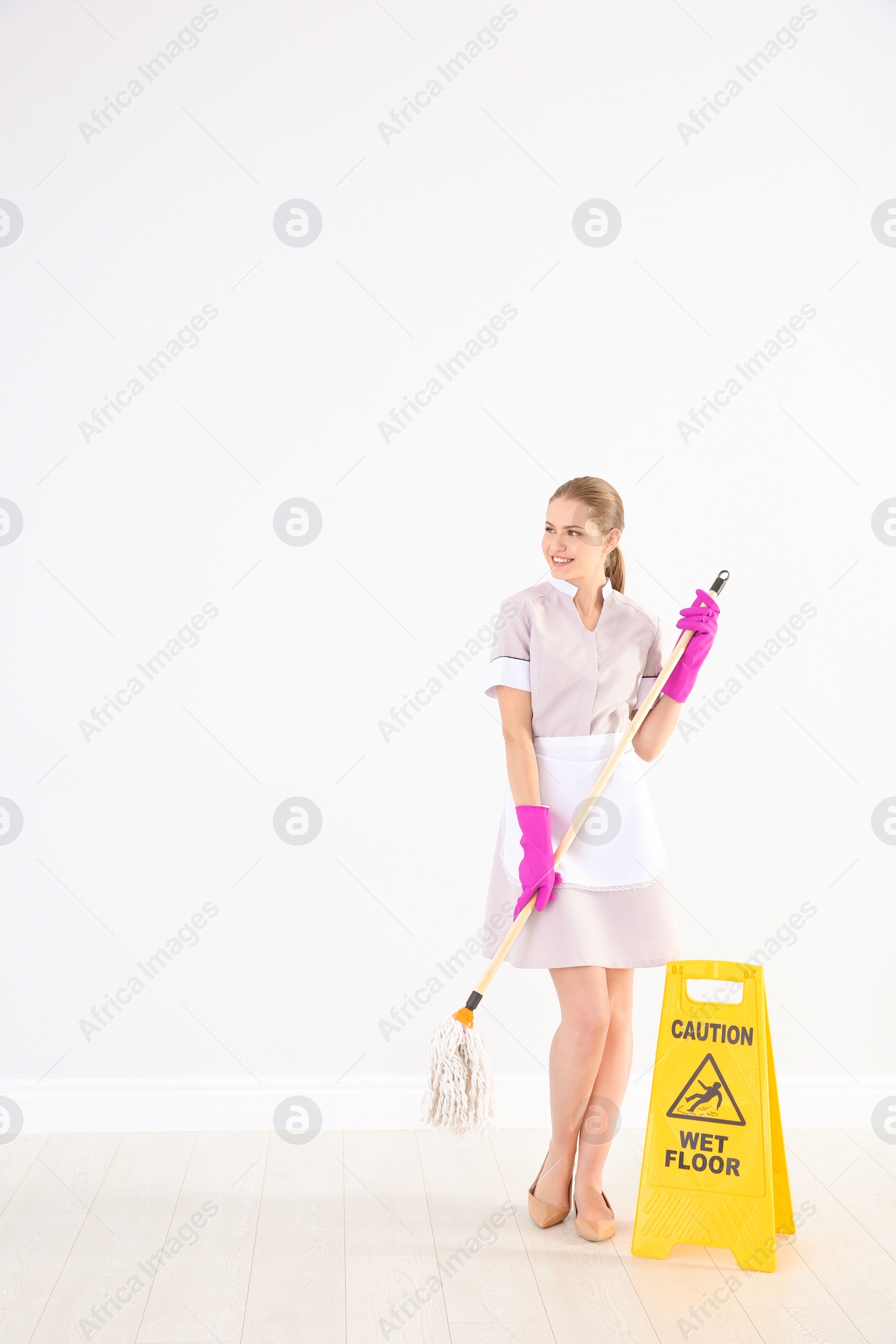 Photo of Young chambermaid with mop near WET FLOOR sign indoors