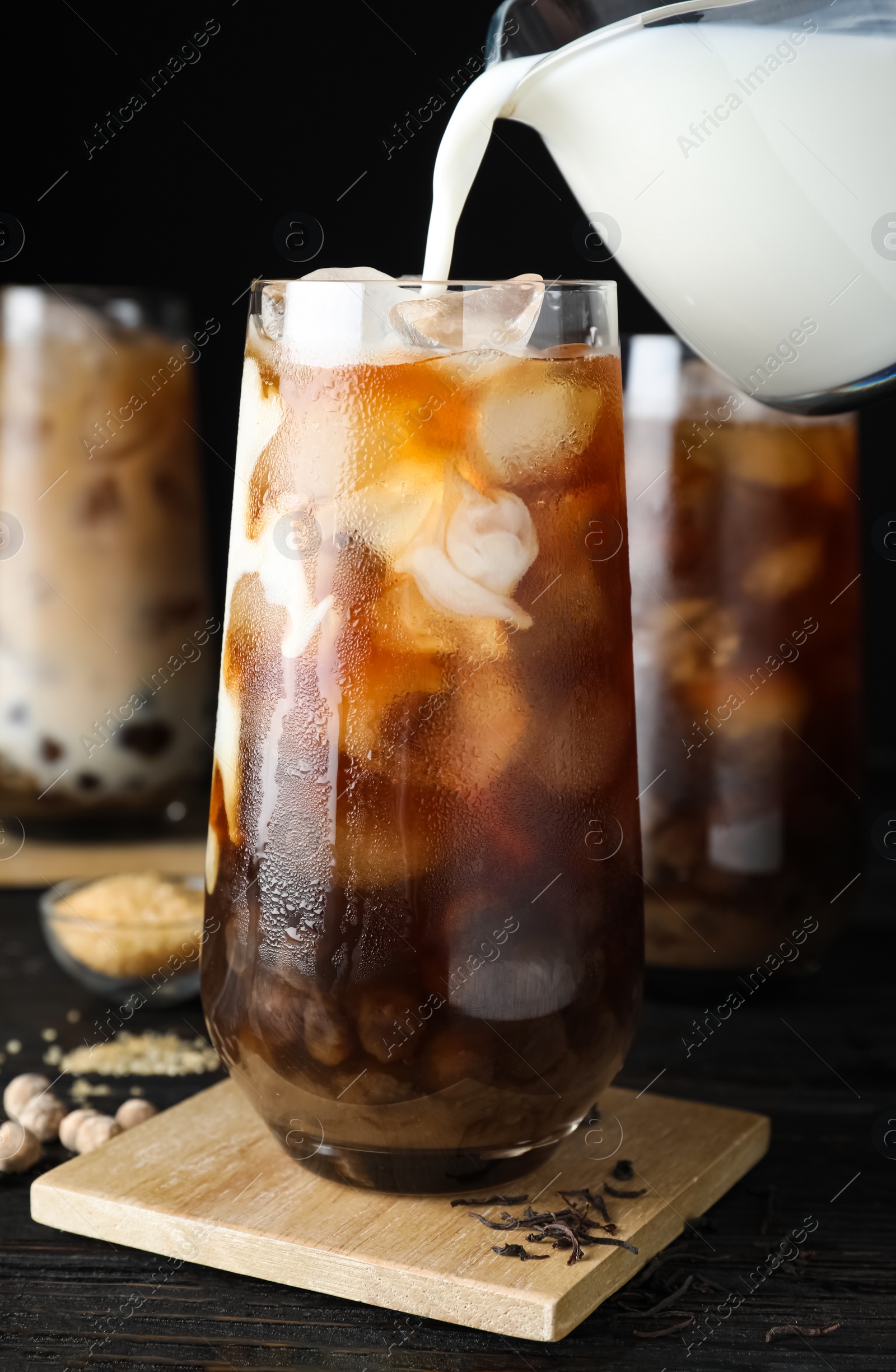 Photo of Pouring milk into glass with bubble tea on black wooden table, closeup