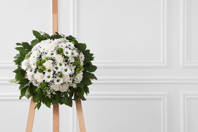 Photo of Funeral wreath of flowers on wooden stand near white wall indoors. Space for text