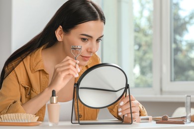 Photo of Beautiful young woman using eyelash curler while doing makeup at table indoors