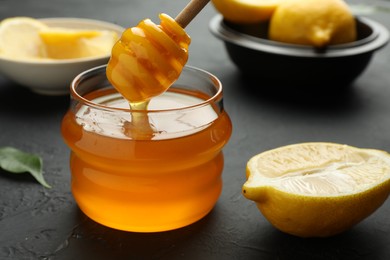Photo of Dripping sweet honey from dipper into jar and fresh lemons on black table, closeup