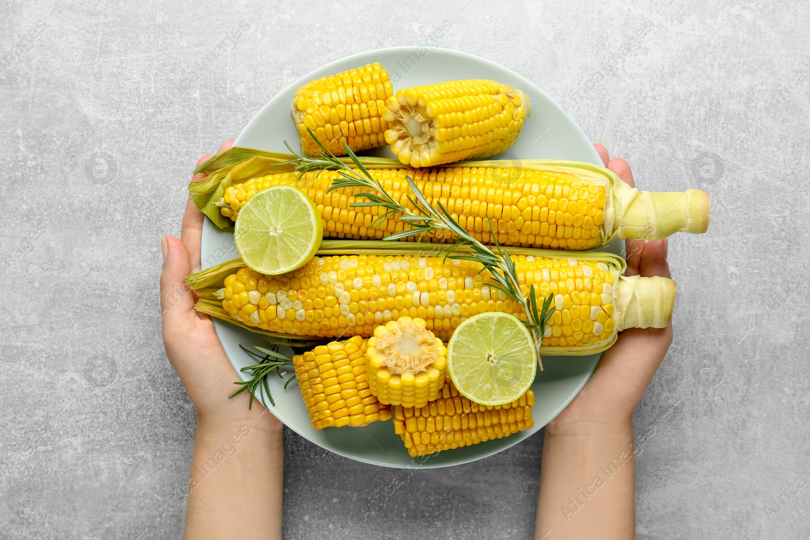 Photo of Woman holding plate with tasty boiled corn at light grey table, top view