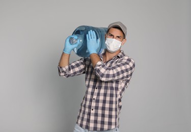 Photo of Courier in medical mask holding bottle for water cooler on light grey background. Delivery during coronavirus quarantine