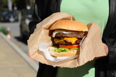 Photo of Woman holding delicious burger in paper wrap outdoors, closeup