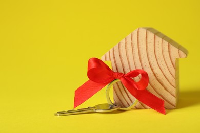 Wooden house model with red bow and key on yellow background, space for text. Housewarming party