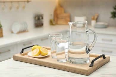 Jug, glasses with clear water and lemons on white table in kitchen