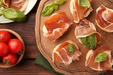 Photo of Board of tasty sandwiches with cured ham and basil leaves near tomatoes on wooden table, flat lay