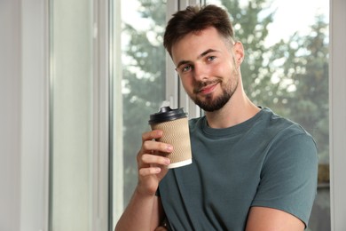 Handsome man with cup of coffee near window indoors, space for text