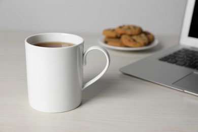 One ceramic mug with drink, laptop and tasty biscuits on white wooden table