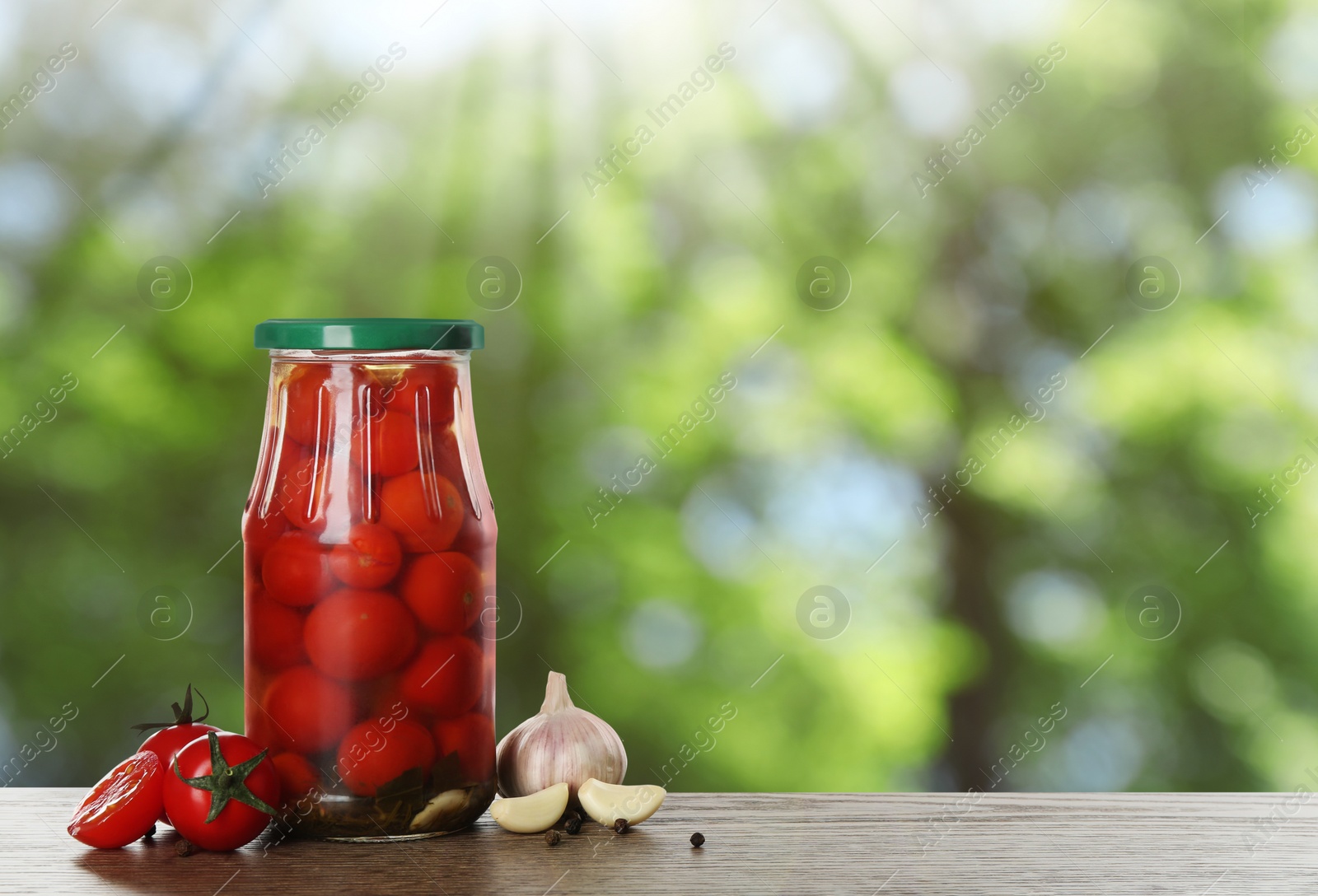 Image of Jar of tasty pickled cherry tomatoes on wooden table against blurred green background. Space for text