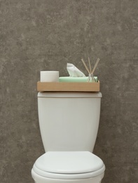 Tray with roll of paper, napkins and reed air freshener on toilet tank near grey wall. Bathroom interior