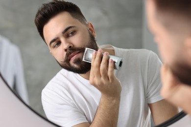 Handsome young man trimming beard near mirror in bathroom