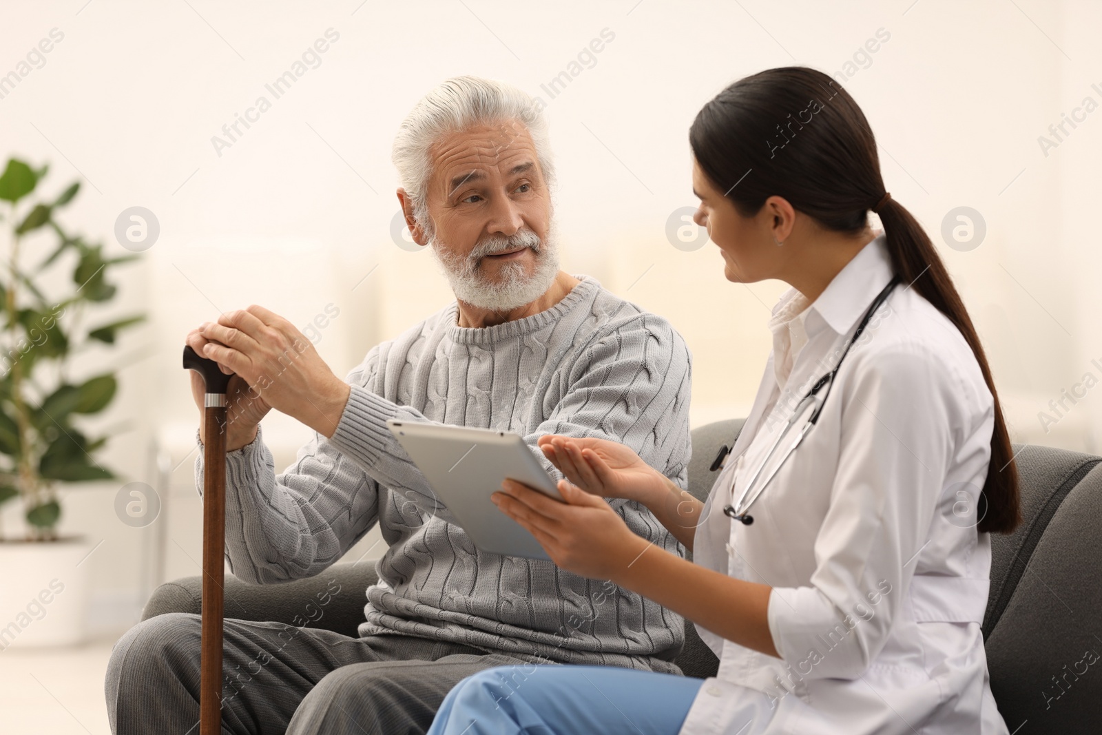 Photo of Smiling nurse with tablet and elderly patient on sofa in hospital