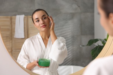 Young woman holding jar of aloe gel near mirror in bathroom