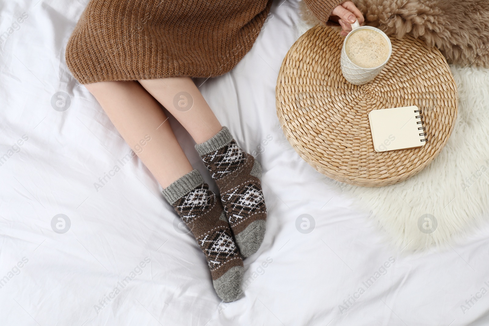 Photo of Woman with cup of hot drink wearing knitted socks on white fabric, top view. Warm clothes