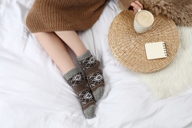 Woman with cup of hot drink wearing knitted socks on white fabric, top view. Warm clothes