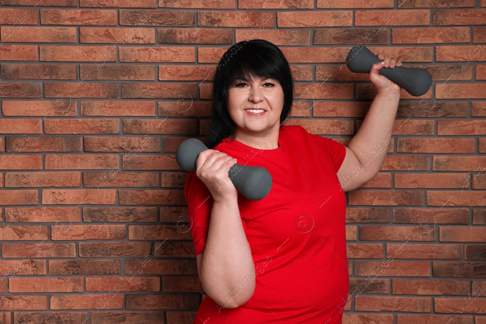 Photo of Happy overweight mature woman doing exercise with dumbbells near brick wall