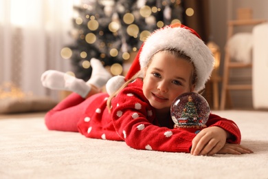 Photo of Little girl in Santa hat playing with snow globe on floor