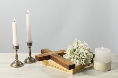 Burning church candles, wooden cross, Bible and flowers on white table