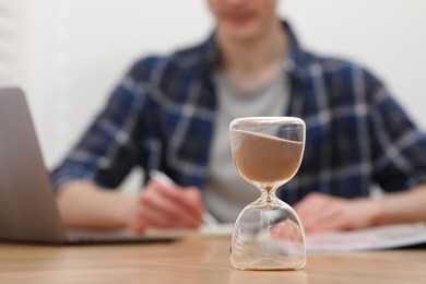 Photo of Hourglass with flowing sand on desk. Man taking notes indoors, selective focus