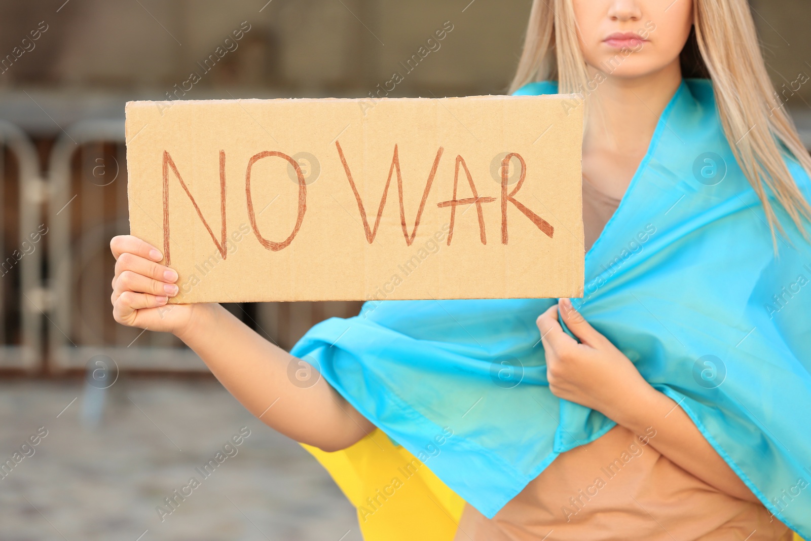 Photo of Sad woman wrapped in Ukrainian flag holding poster with words No War against blurred background, closeup