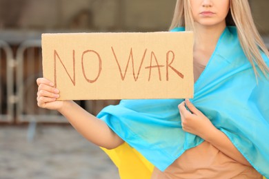 Photo of Sad woman wrapped in Ukrainian flag holding poster with words No War against blurred background, closeup