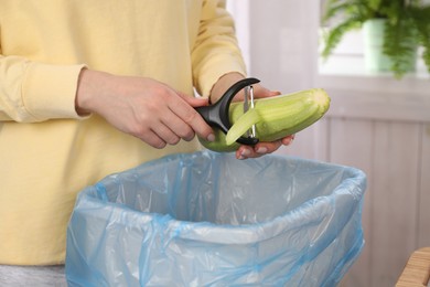 Photo of Woman peeling fresh zucchini above garbage bin indoors, closeup