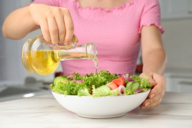 Photo of Woman pouring oil from jug into bowl with salad in kitchen, closeup