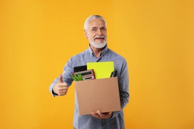 Photo of Happy unemployed senior man with box of personal office belongings showing thumb up on orange background