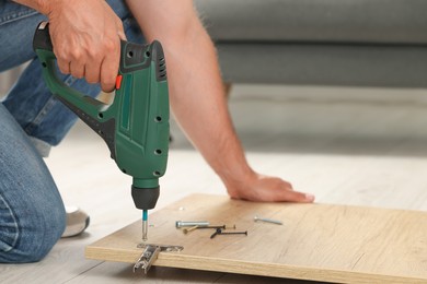 Photo of Man with electric screwdriver assembling furniture on floor indoors, closeup