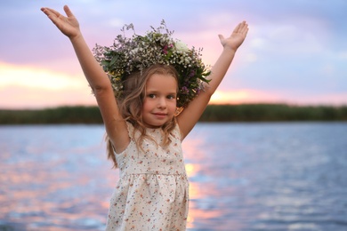 Photo of Cute little girl wearing wreath made of beautiful flowers near river at sunset